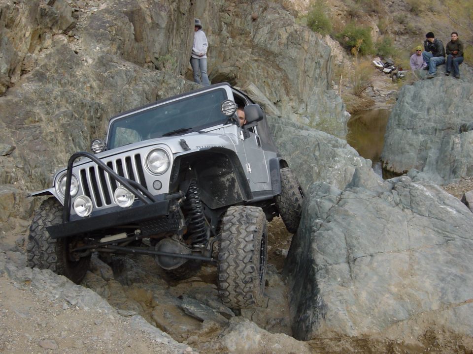 Silver Jeep Wrangler TJ with aftermarket bumper and lift kit crawling over large rocks