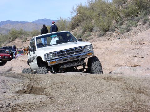 1986 Toyota Hilux - A white Toyota pickup truck is shown off-roading on a dirt path with its front passenger side wheel significantly elevated.