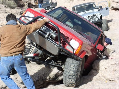 1986 Toyota Hilux - A red Jeep Cherokee XJ is stuck on rocks with people trying to help, while a silver Jeep Wrangler waits behind.
