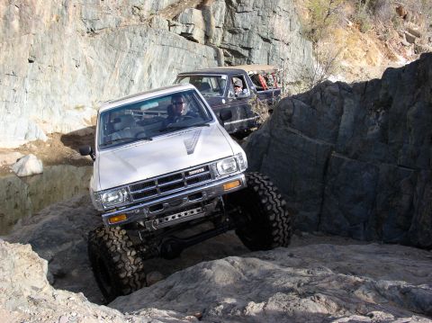 1986 Toyota Hilux - Two trucks navigate a challenging rocky trail, with the lead vehicle showing significant off-road modifications.