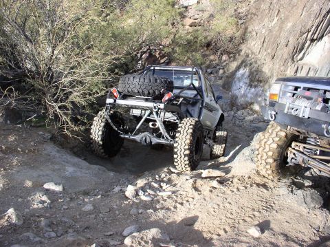 1986 Toyota Hilux - Two modified pickup trucks tackle a challenging off-road course on a sunny day.