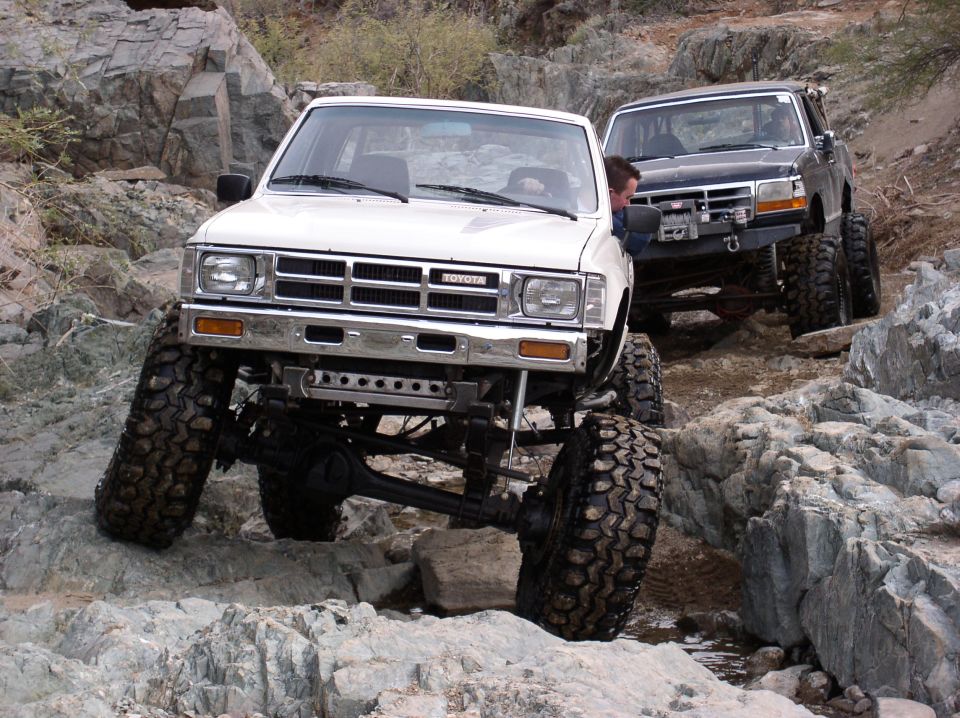 White 1986 Toyota Hilux and black Ford Bronco off-roading on rocks