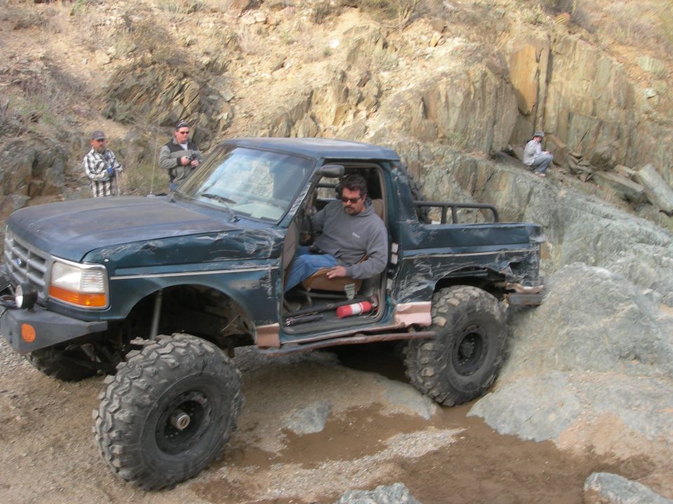 Dark green Ford pickup truck with large off-road tires and a damaged body, seen from the front-left