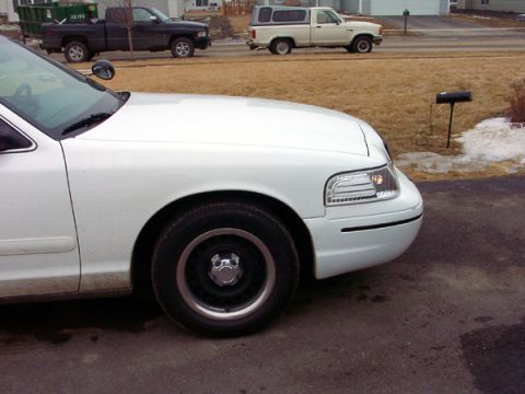 1999 Ford Crown Victoria - Front quarter panel and wheel of a white sedan, with other vehicles visible in the background.