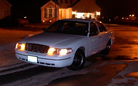 1999 Ford Crown Victoria - A white sedan is parked outside a house at night, with its headlights illuminated.