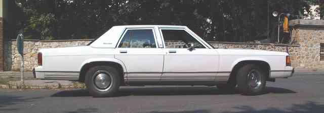 White full-size four-door sedan viewed from driver's side, parked by stone wall.