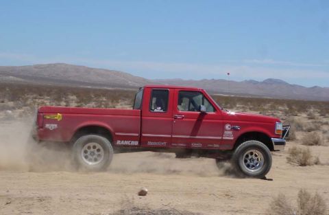 1996 Ford Bronco - A red pickup truck drives off-road, creating a cloud of dust under a clear blue sky.