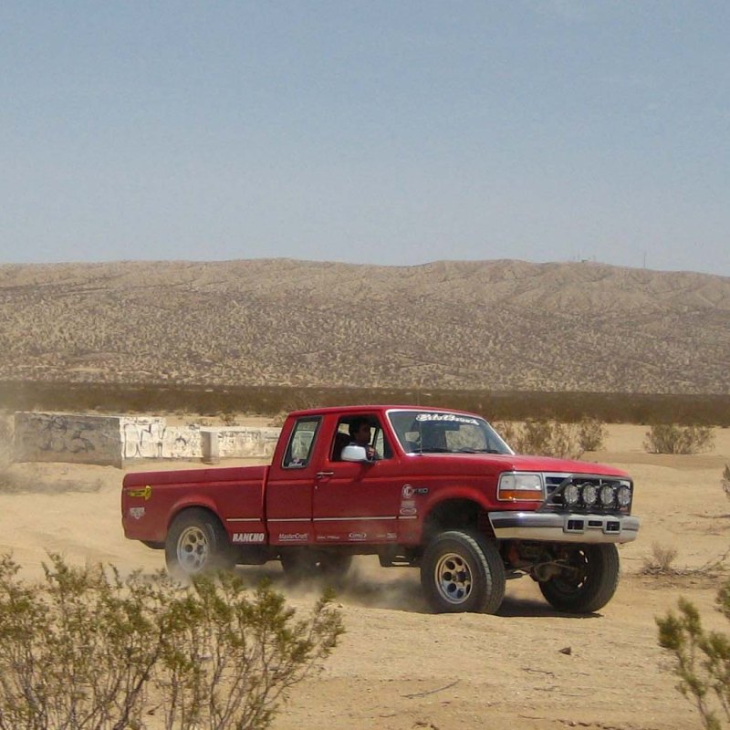 Red 1995 Ford F-250 extended cab pickup truck with off-road lights driving on a dirt road in a desert landscape