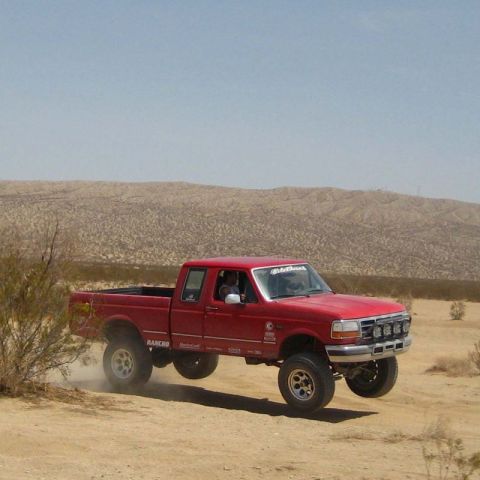 1996 Ford Bronco - A red pickup truck navigates a dusty desert terrain with its front wheels off the ground.