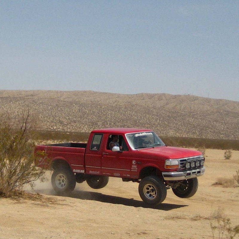 Red pickup truck with lifted suspension driving off-road in a desert landscape. It has a crew cab and a long bed