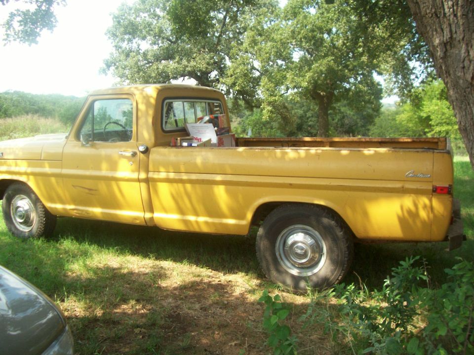 Driver-side profile of a yellow vintage Ford Custom pickup in dappled shade