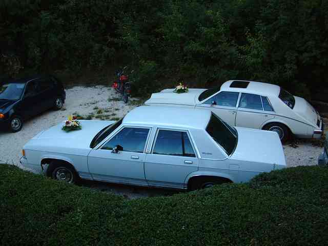 White four-door sedan parked on gravel with flowers on hood, other cars behind