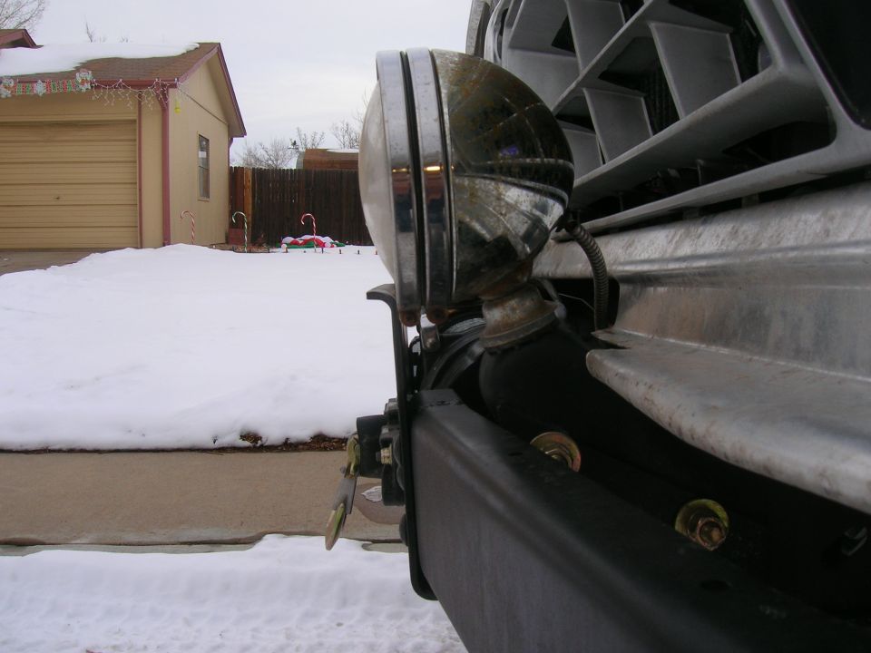 Chrome round off-road driving light on a black front bumper, snowy driveway in background