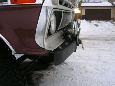 1979 Ford Bronco - Low close-up of the front corner of a maroon and white Ford truck parked on snow, showing the FORD-lettered white grille panel, single roun…