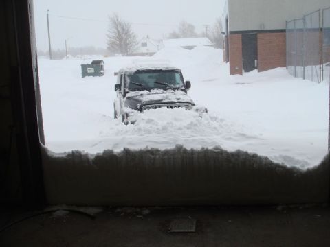 1996 Ford Bronco - A black Jeep Wrangler is partially buried in a large snowdrift, viewed from inside a garage.