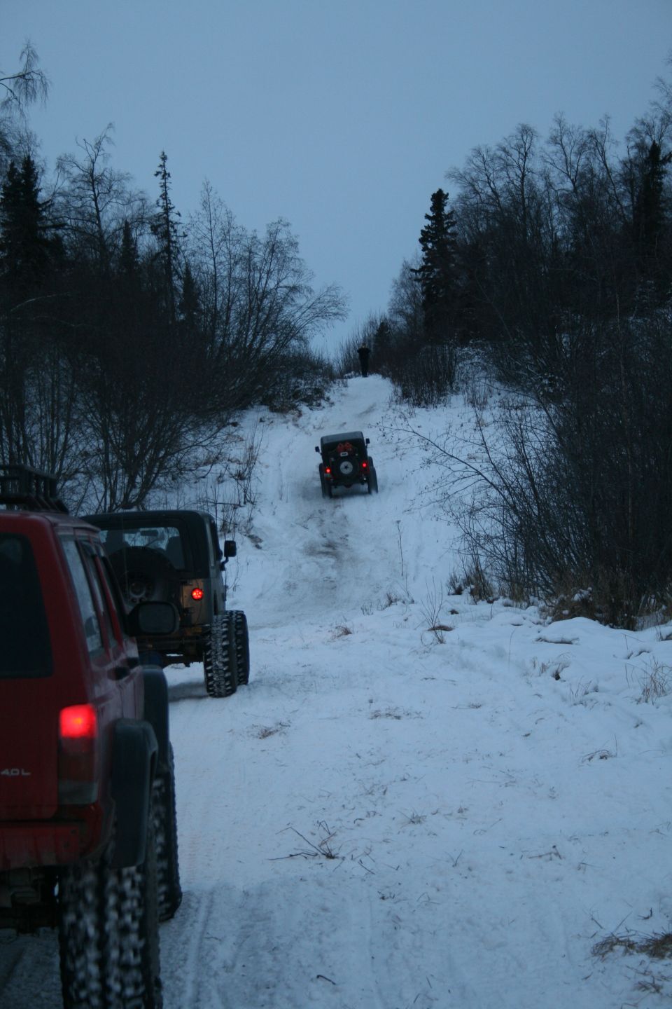 Three Jeeps driving up a snow-covered hill in a wooded area. The lead vehicle is red, followed by two dark-colored Jeeps