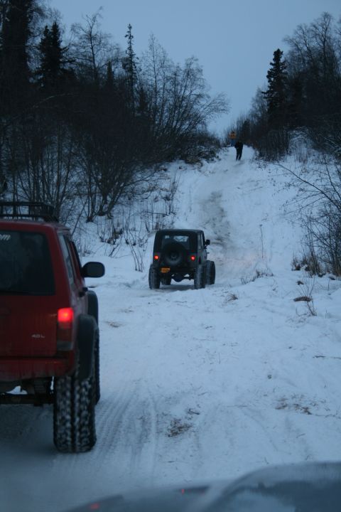 2003 Jeep TJ - Two Jeeps navigate a snowy trail with trees on either side and a person visible further up the incline.