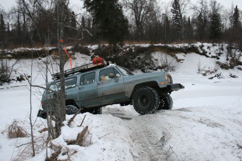 2003 Jeep TJ - A modified SUV with oversized tires drives through a snowy, off-road environment near a frozen body of water.