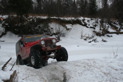 2003 Jeep TJ - A red 2003 Jeep TJ navigates a snowy embankment near a frozen body of water.
