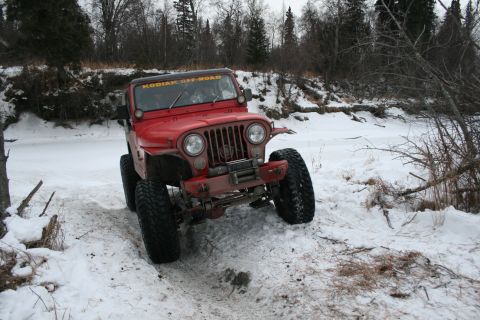 2003 Jeep TJ - Red Jeep CJ-7 navigates a snowy path near a frozen river, trees in background.