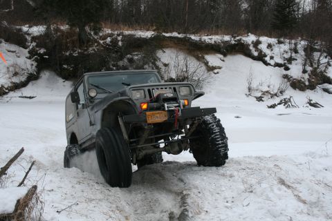 2003 Jeep TJ - A gray Jeep Wrangler navigates a snowy terrain, its large tires kicking up snow as it moves forward.
