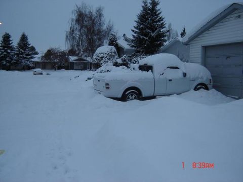 1990 Ford Bronco - A white pickup truck is almost completely covered in snow, with only the roof and part of the side visible. A garage is to the right, also…