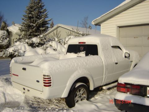 1990 Ford Bronco - A white pickup truck sits covered in snow next to a residential garage on a clear winter day.
