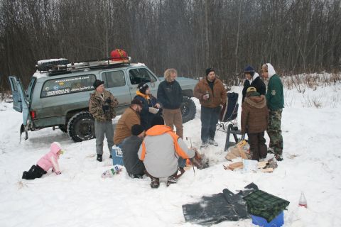 2003 Jeep TJ - A group of people and a child are gathered around a campfire in a snowy wooded area next to a Chevrolet Suburban.