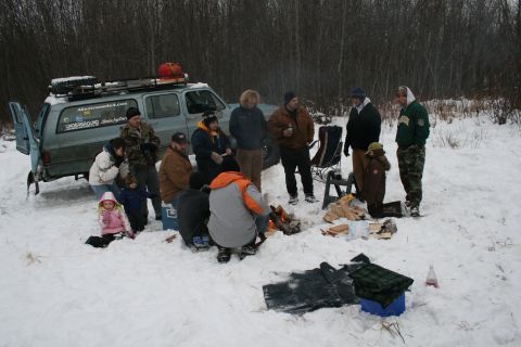 2003 Jeep TJ - A group of people gather around a campfire next to a light blue SUV in a snowy, wooded area.