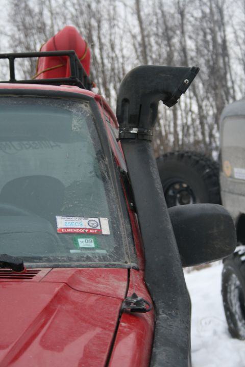2003 Jeep TJ - A red off-road vehicle is equipped with a snorkel and a roof rack, parked in a snowy environment with another vehicle nearby.