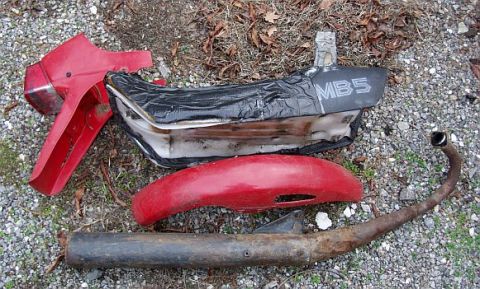 1985 Ford Crown Victoria LTD - Scattered red motorcycle parts, including a seat labeled MB5, fender, exhaust pipe, and tail section, lie on a gravel surface.
