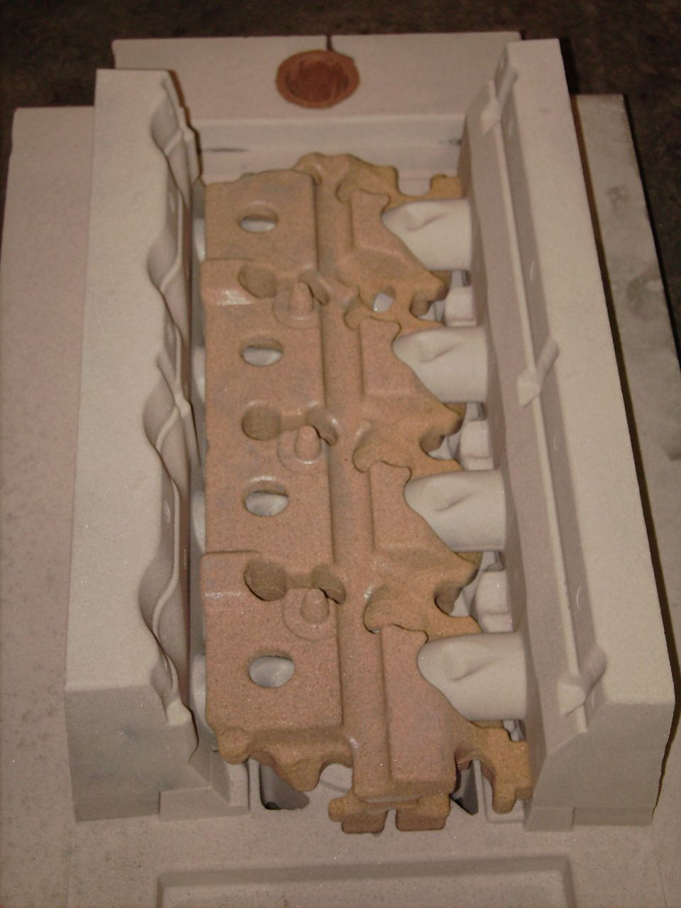 Tan sand-cast cylinder head core in white foam box with penny for scale