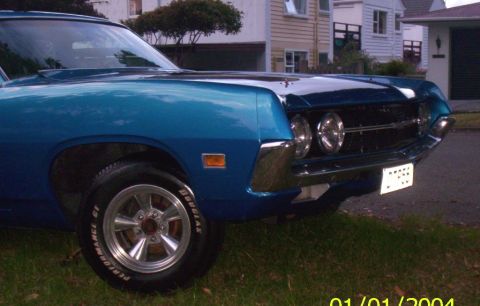 1970 Ford Ranchero - A blue coupe is parked on grass in front of houses, showing its front quarter panel and grille.