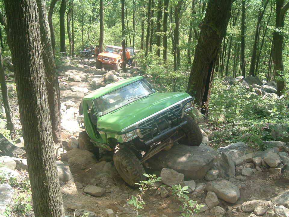 1978 Ford Bronco climbing over large rocks on a wooded trail; another orange vehicle is visible further up the path