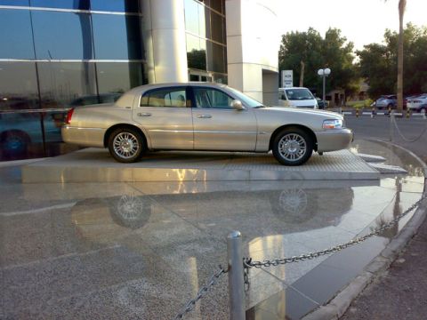 1999 Mercury Grand Marquis - A gold Lincoln Town Car is parked on a raised platform, reflecting in the wet ground below.