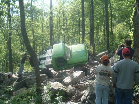 1978 Ford Bronco - A green off-road vehicle is overturned on a rocky trail in a wooded area, with onlookers watching.