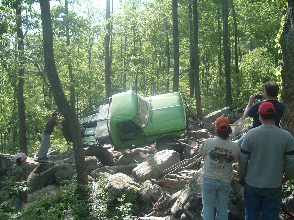 Green off-road vehicle tipped on its side amidst rocks and trees, with people observing