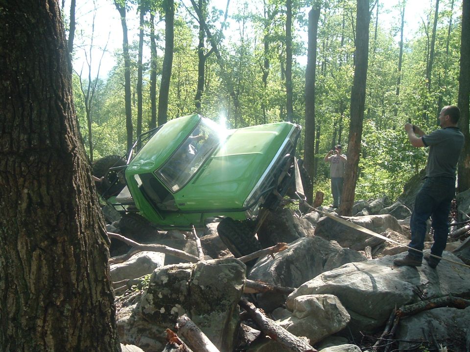 Green off-road vehicle tipped over on rocks in woods, two people taking photos