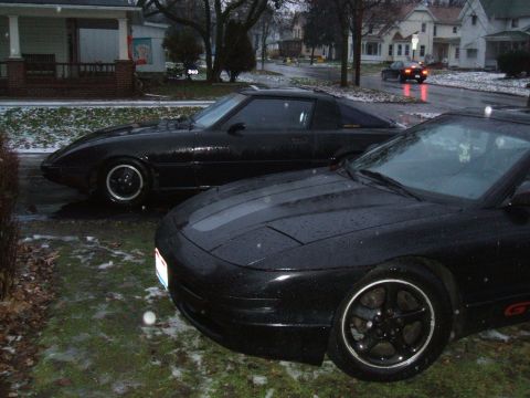 1983 Mazda RX-7 - Two black cars, a Mazda RX-7 and a Ford Probe, are parked on a wet driveway.