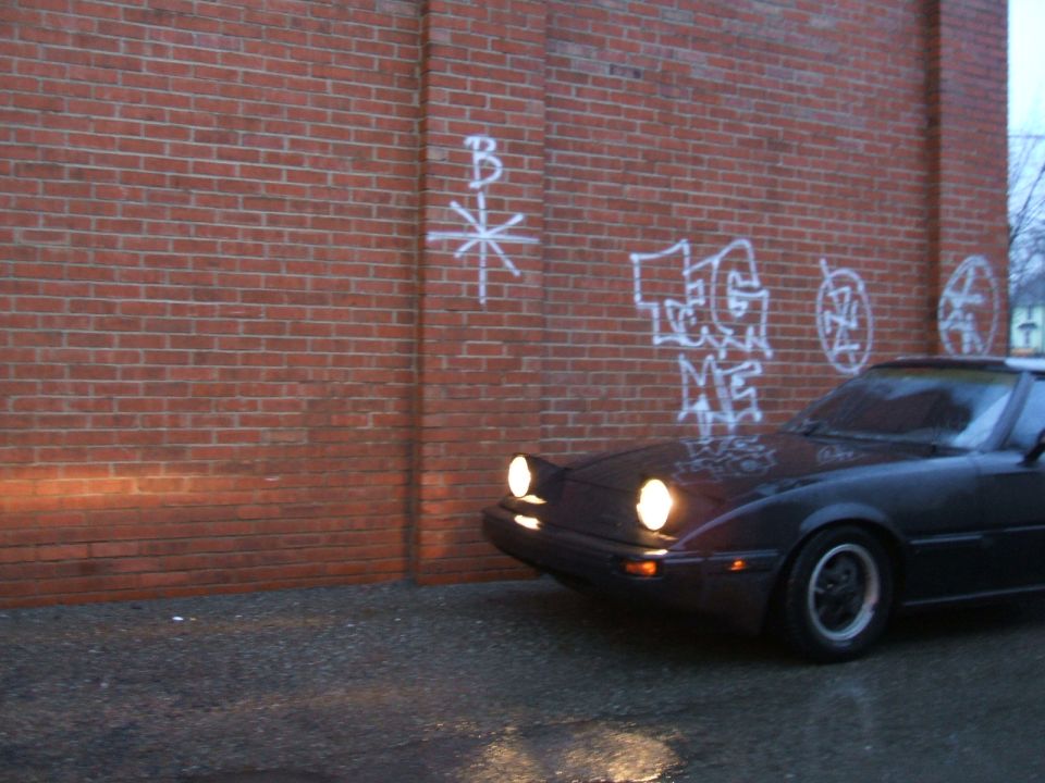 Dark coupe with pop-up headlights parked near a brick wall with graffiti