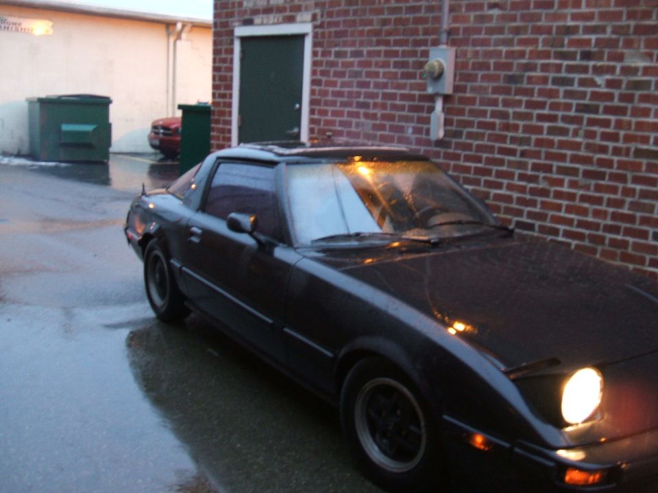 1983 Mazda RX-7 parked on wet asphalt near a brick building, front passenger side visible with headlight on