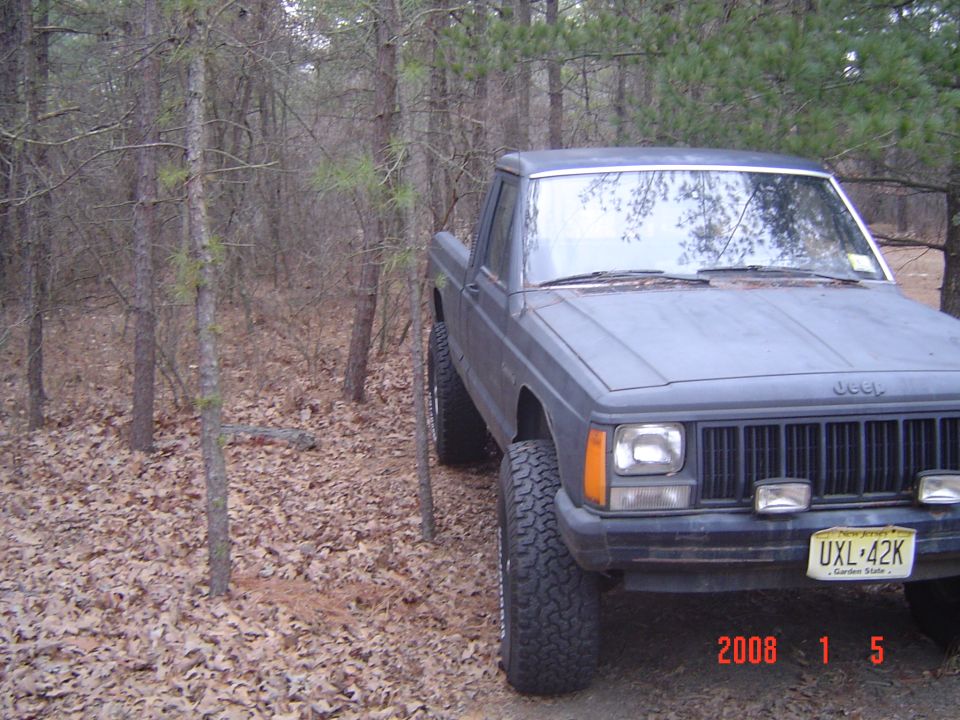 1988 Jeep Comanche pickup truck parked on a leaf-covered ground in a wooded area. Front grille and headlights visible