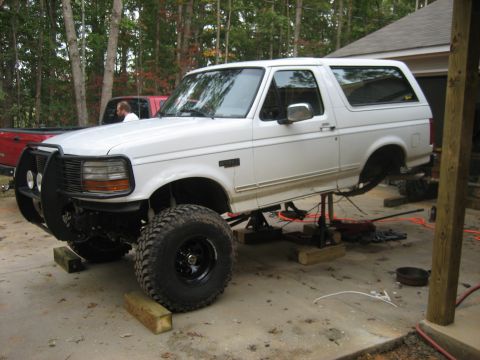 1996 Ford Bronco - White full-size Ford Bronco raised on jack stands in a driveway garage, fitted with a brush guard, KC-style lights and large mud-terrain ti…