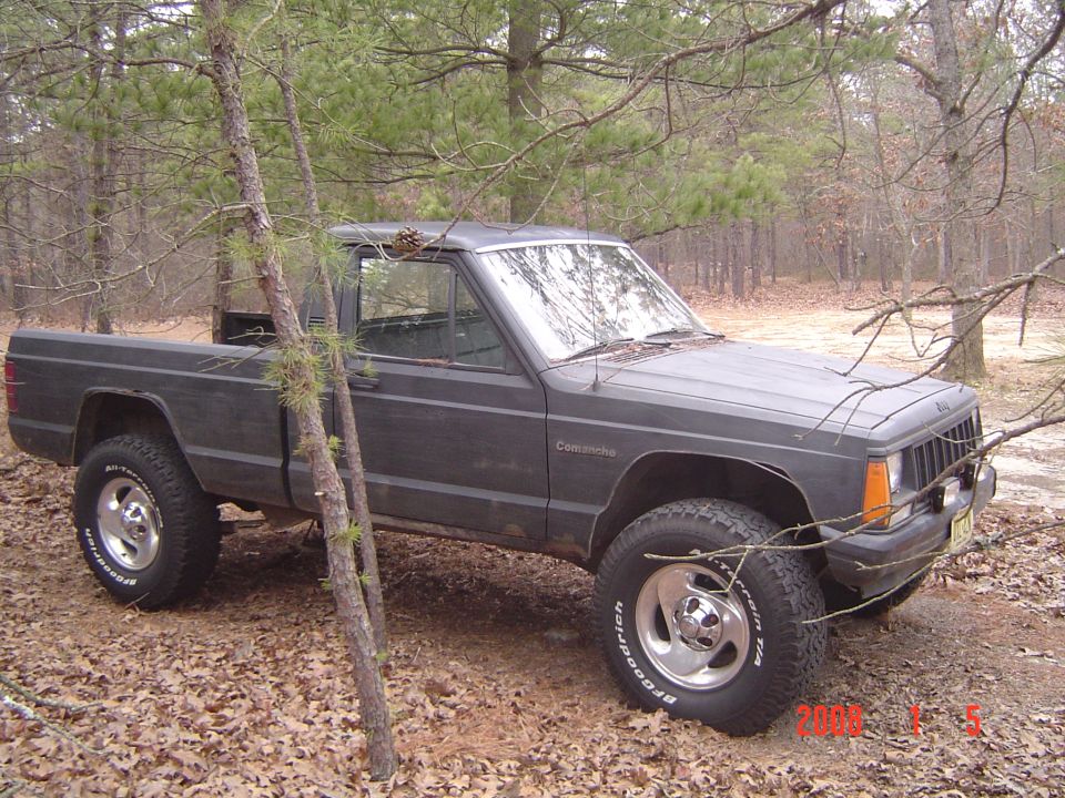Flat-black Jeep Comanche pickup parked in pine woods on fallen leaves