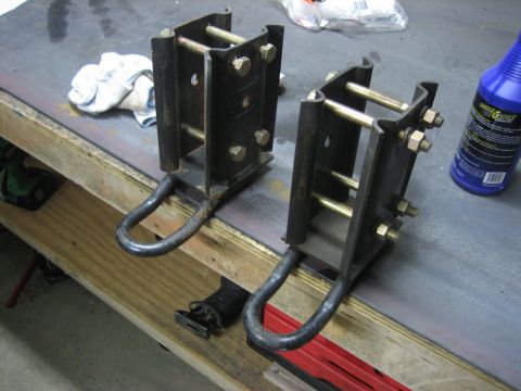 1996 Ford Bronco - A pair of black steel leaf-spring shackle assemblies with large U-bolts and yellow-zinc hardware sit on a workbench, one taller than the ot…