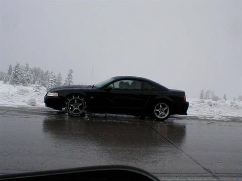 2003 Ford Crown Victoria - A black coupe is seen from the side in a snowy environment, with snow chains fitted to the front wheel.