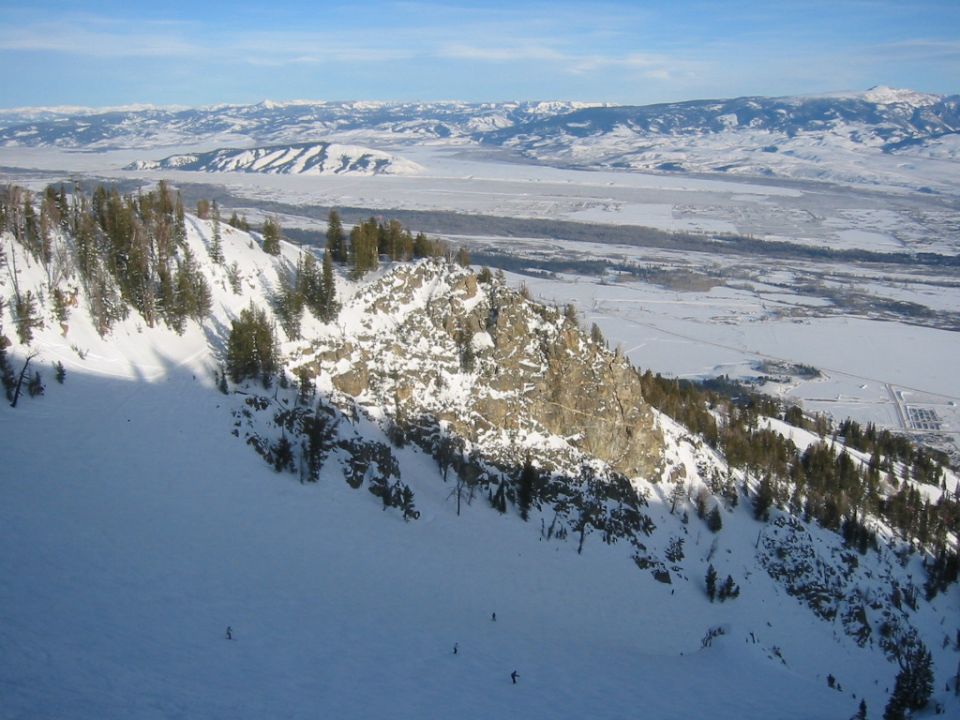 Snowy mountain landscape with skiers on a slope, distant valley and mountains under a blue sky. Supermotors.net logo in corner