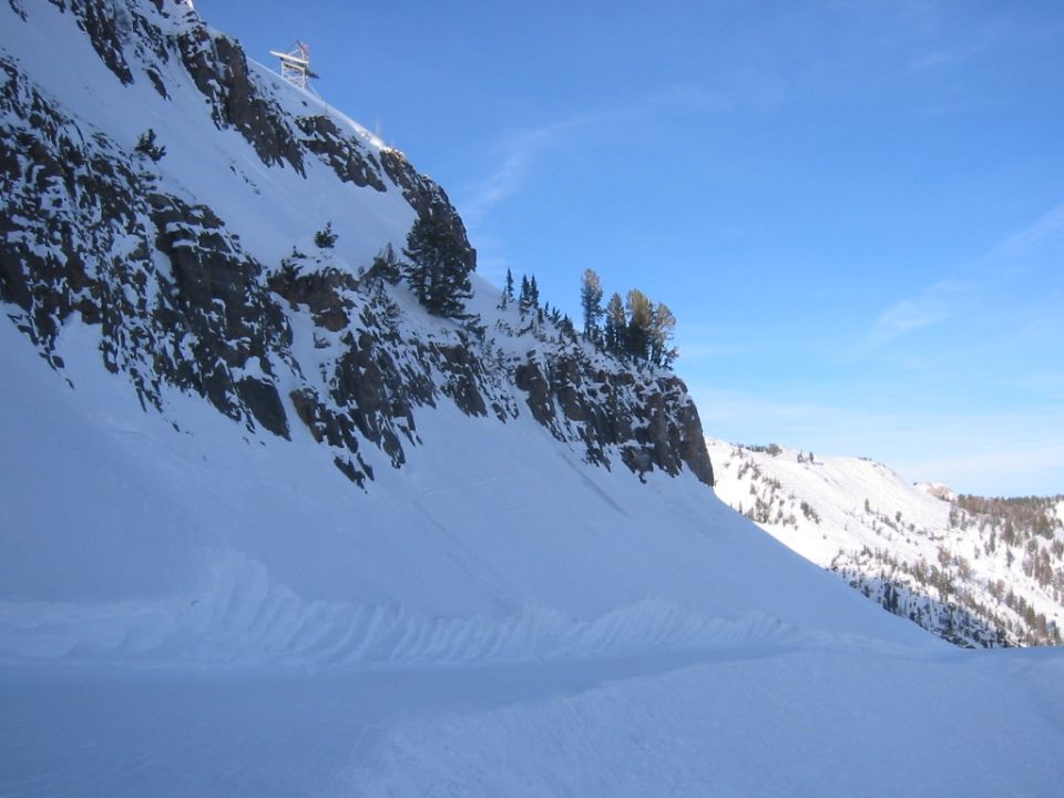 Snowy mountain slope with trees and a ski lift tower against a clear blue sky. Supermotors.net logo in the corner