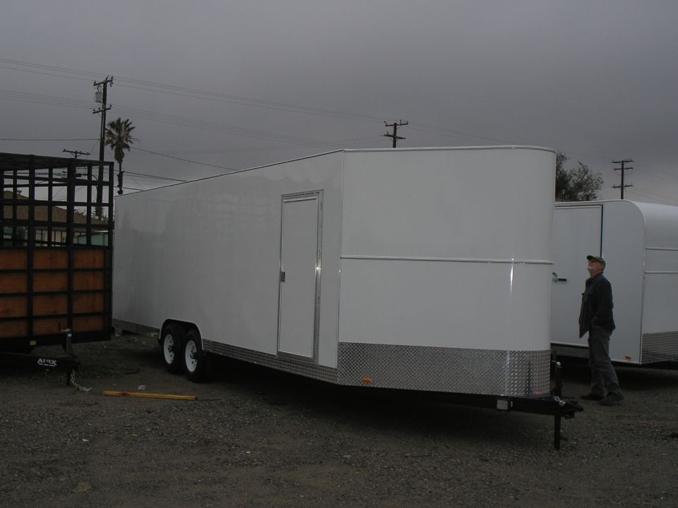 Enclosed white trailer with a side door, parked next to a black cage trailer. A man stands near the trailer door