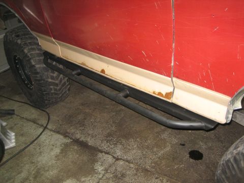 1990 Ford Bronco - Aftermarket rock sliders are installed on the side of a red vehicle, showing wear on the tan trim.